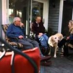 Three individuals, including a veteran and caregivers, relax on a porch with a dog, illustrating the community and companionship aspect of the Medical Foster Home program for veterans.