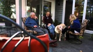 A veteran, caregiver, and another person sit on a porch with a dog, illustrating the community and home-like atmosphere of the veteran foster care program.